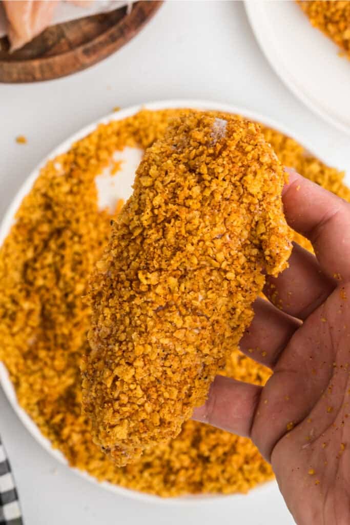 Hand holding a raw chicken tenderloin coated in crushed corn chip mixture.