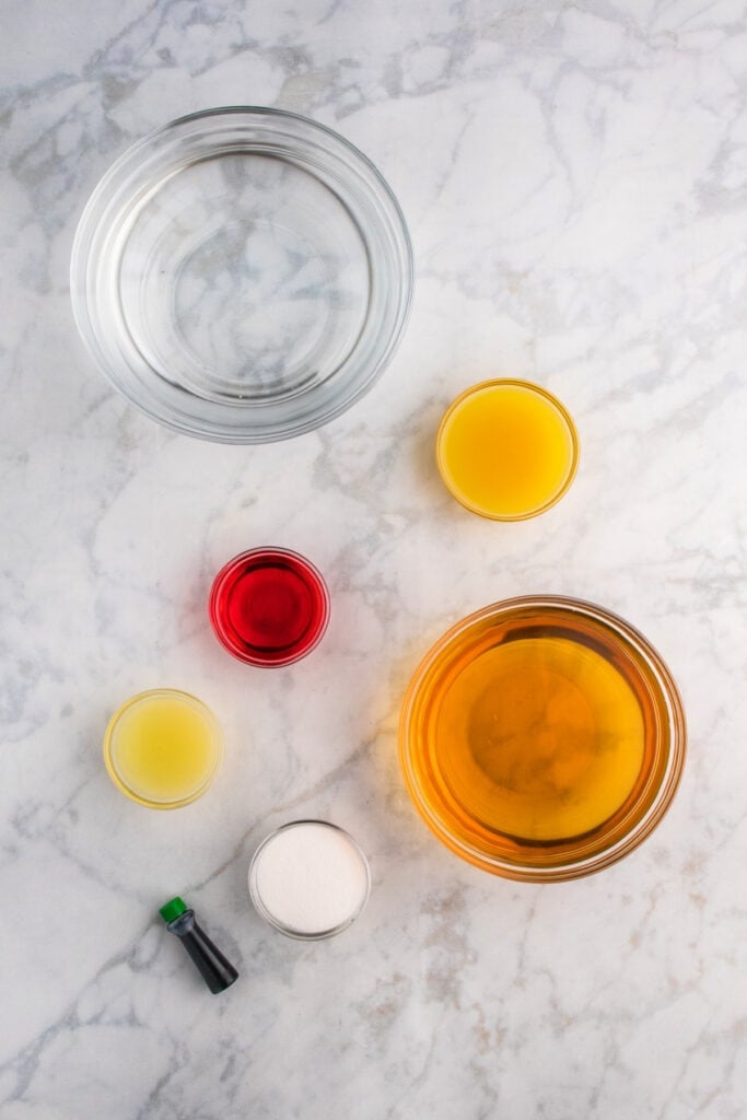 Glass bowls of ingredients including apple juice, pineapple juice, lime juice, grenadine, sugar, and green food coloring on marble surface.