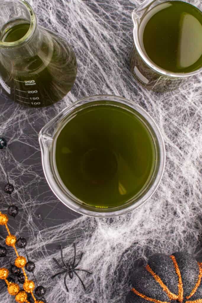 Three glass beakers filled with dark green Toxic Waste Punch on a cobweb-decorated table with Halloween beads and pumpkins.