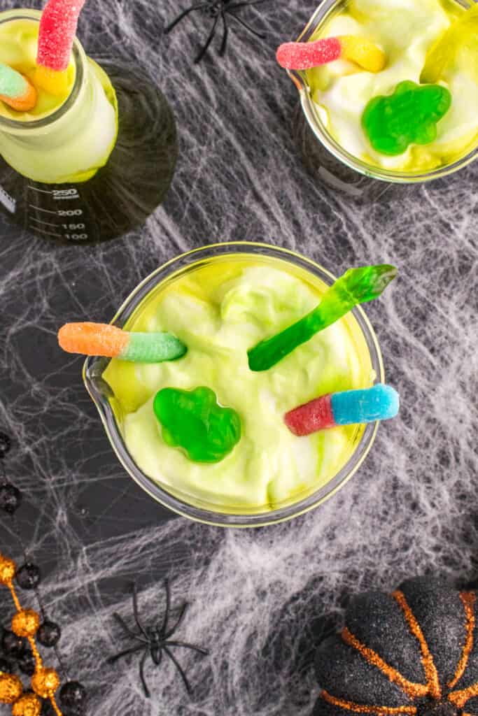 Beakers of Toxic Waste Punch decorated with gummy worms and gummy frogs on foamy green topping for a creepy Halloween drink.