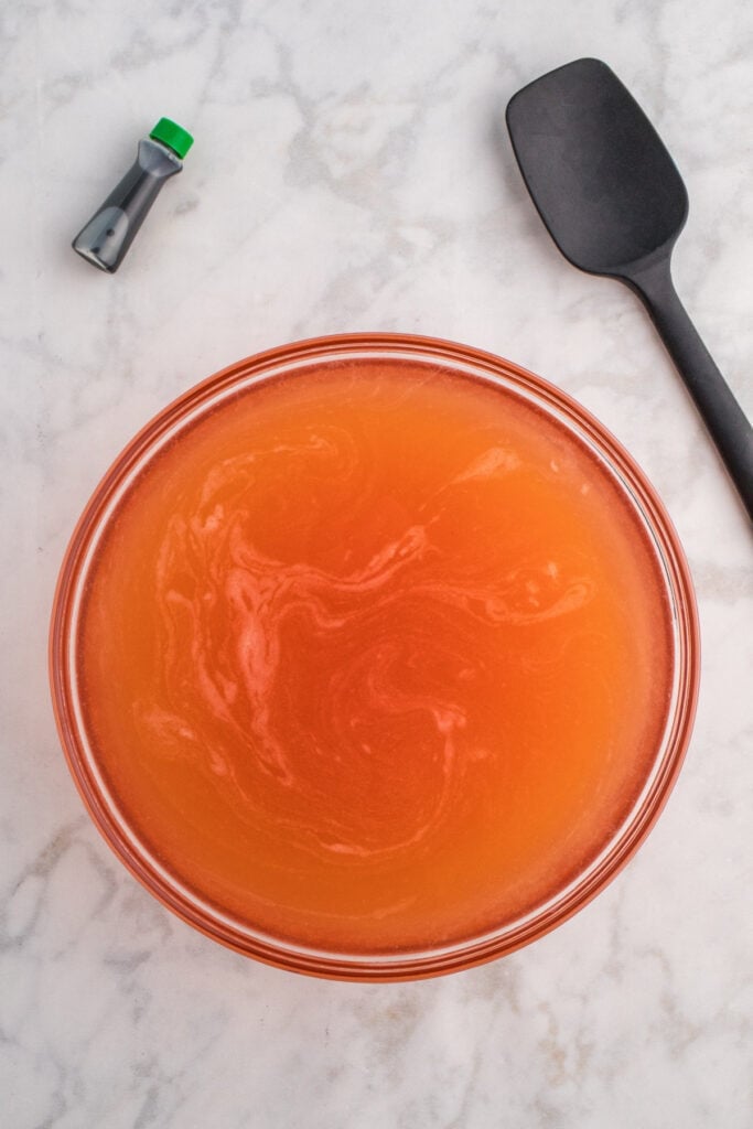 Large glass bowl filled with orange punch mixture and a black spatula on the side with green food coloring bottle nearby.
