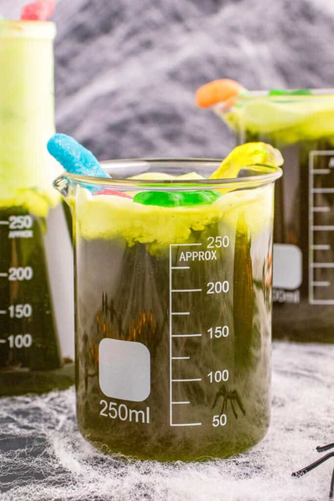 Close-up of a beaker filled with Toxic Waste Punch, topped with green foam and gummy decorations, with cobwebs in the background.