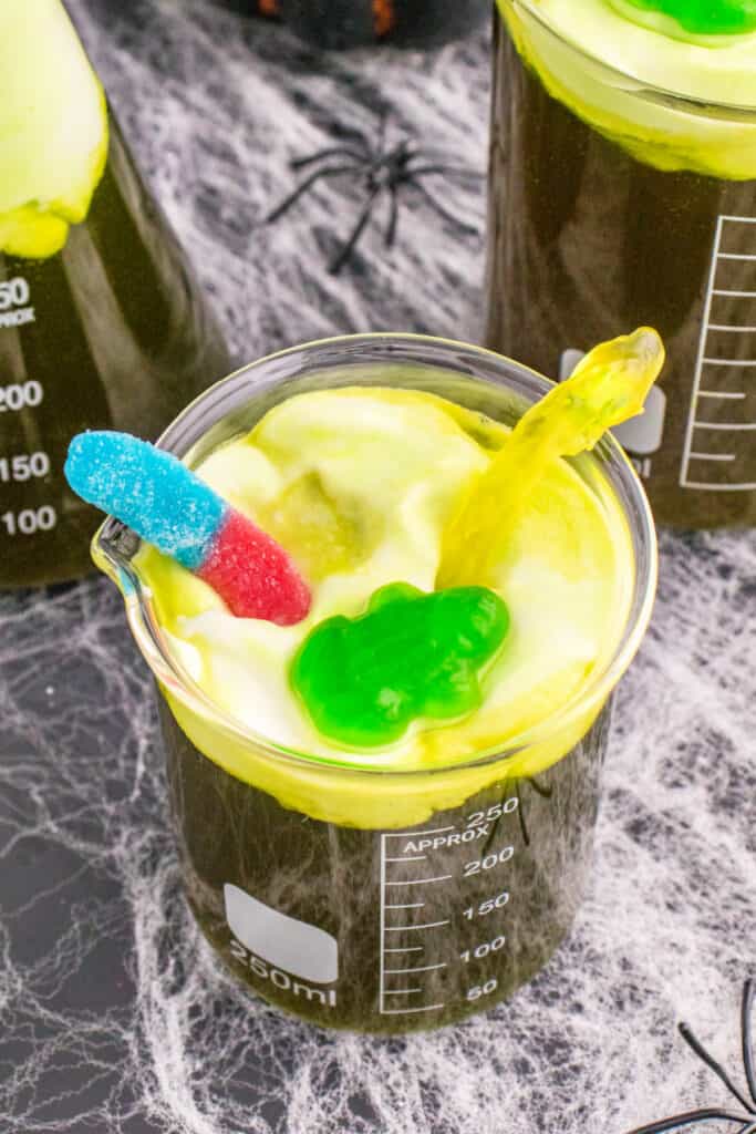 Beaker of Toxic Waste Punch with gummy frog and gummy worm toppings, showcasing the foamy toxic waste effect.