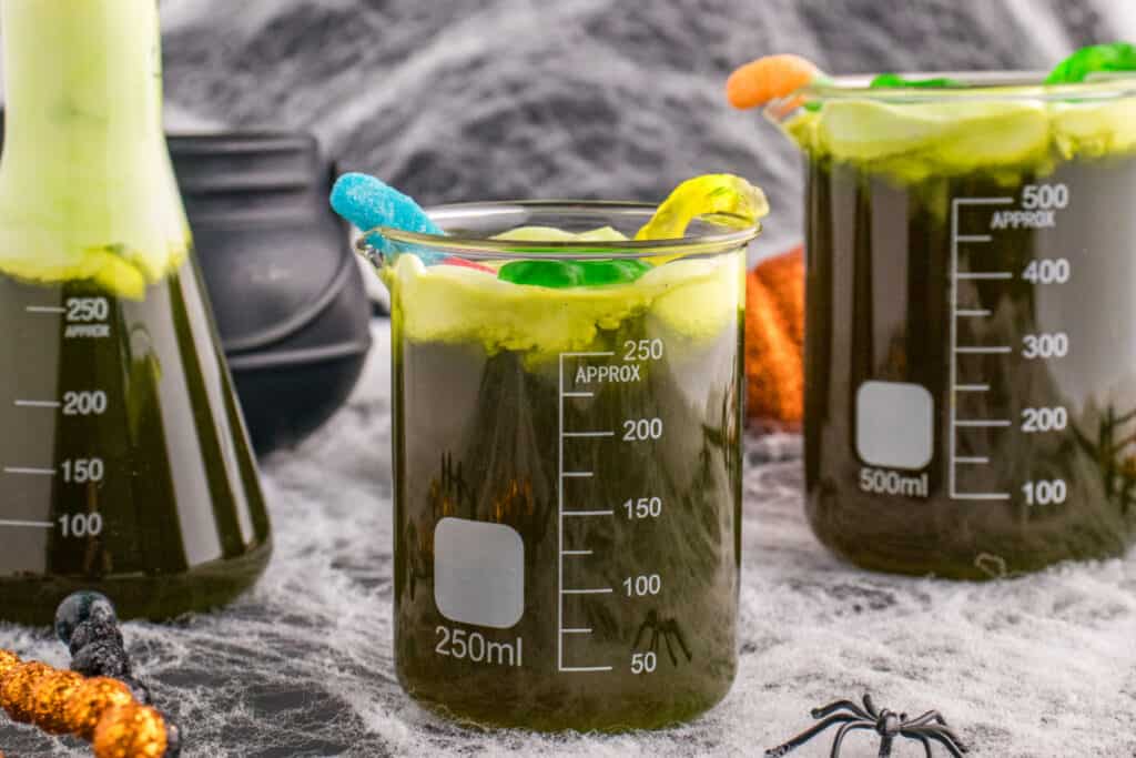 Halloween table with multiple beakers of Toxic Waste Punch topped with green foam and gummy candy, surrounded by cobwebs and spiders.