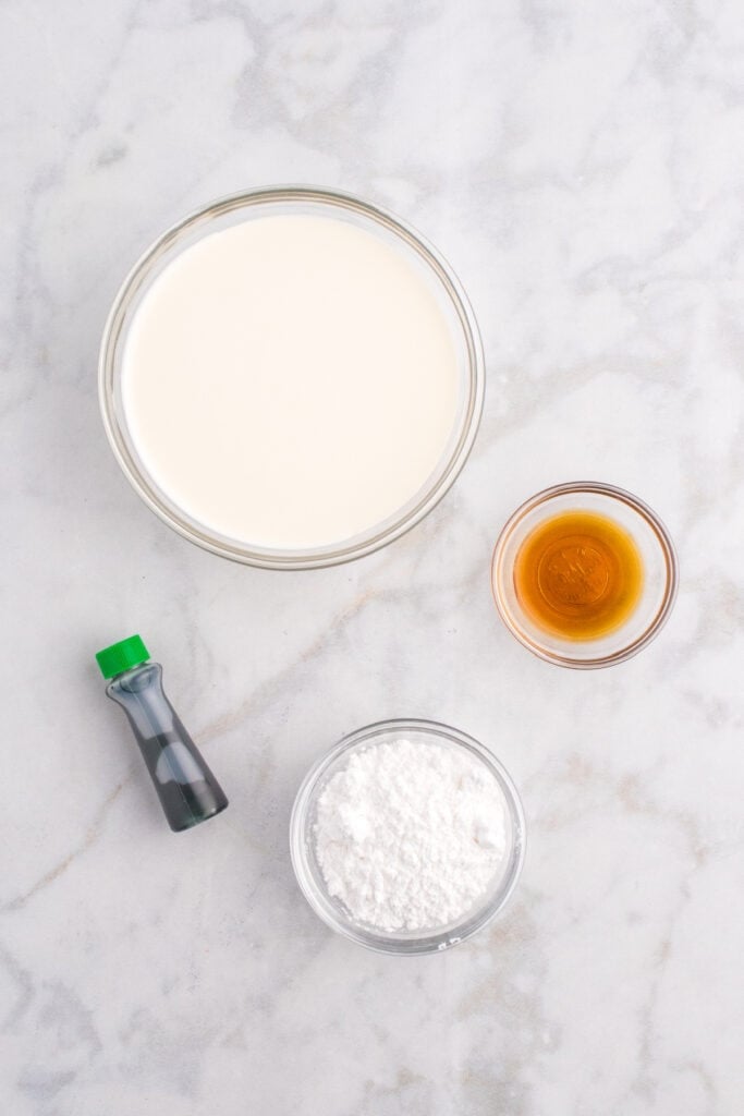 Bowls of heavy cream, powdered sugar, vanilla extract, and green food coloring arranged on a marble countertop.