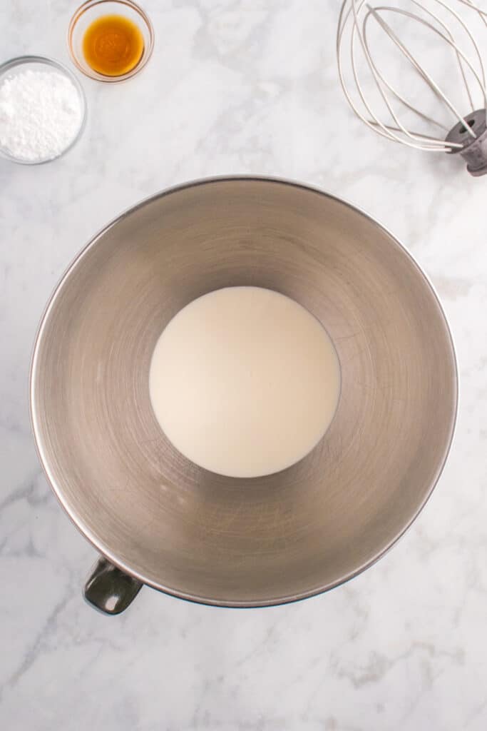 Mixing bowl with heavy cream at the bottom, ready to be whipped with a whisk attachment and vanilla extract nearby.