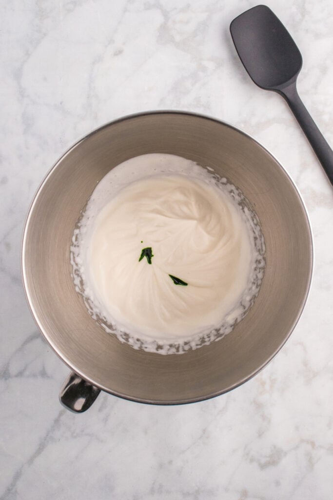 Mixing bowl with whipped cream partially mixed and drops of green food coloring on top before being swirled.