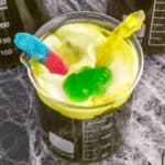 Square cropped image of a beaker filled with Toxic Waste Punch topped with foamy green whipped cream and gummy candy decorations.