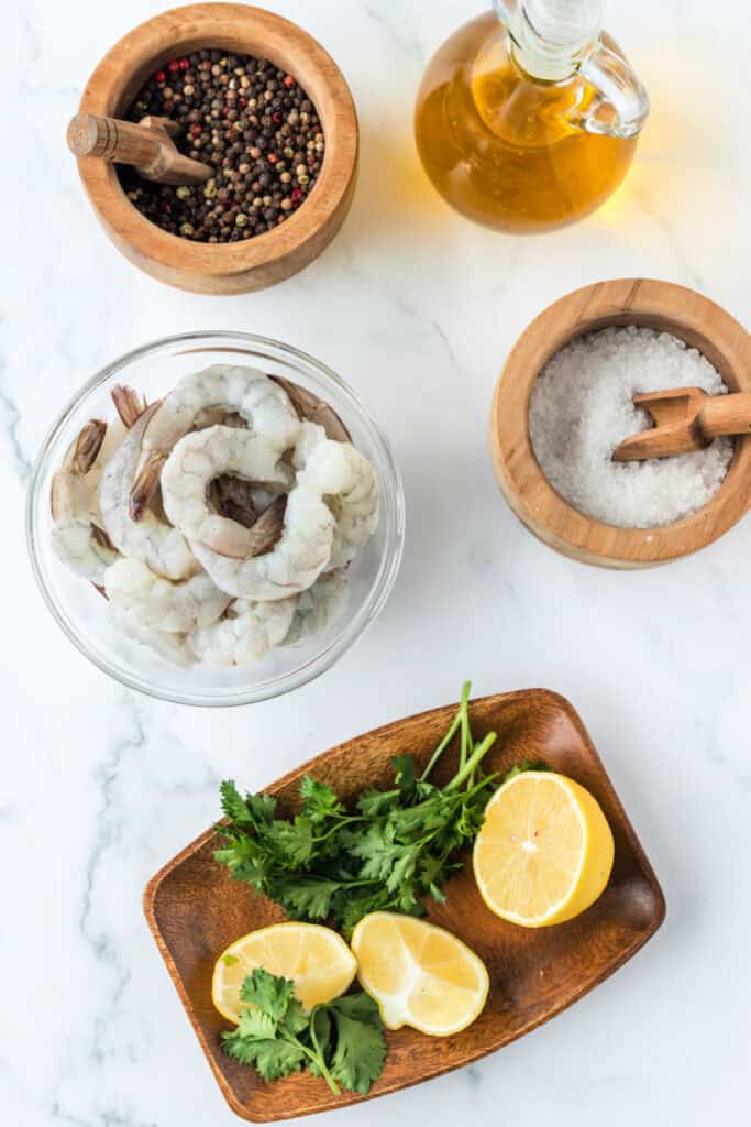 Overhead view of shrimp, lemons, parsley, salt, peppercorns, and oil arranged on a marble surface for Air Fryer Shrimp ingredients.