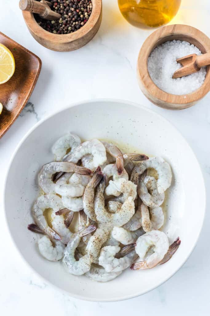 Raw shrimp seasoned in a white bowl surrounded by salt, pepper, lemons, and olive oil for Air Fryer Shrimp preparation.