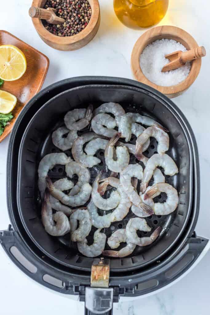 Uncooked shrimp arranged in a single layer in an air fryer basket before cooking, with seasoning ingredients nearby.