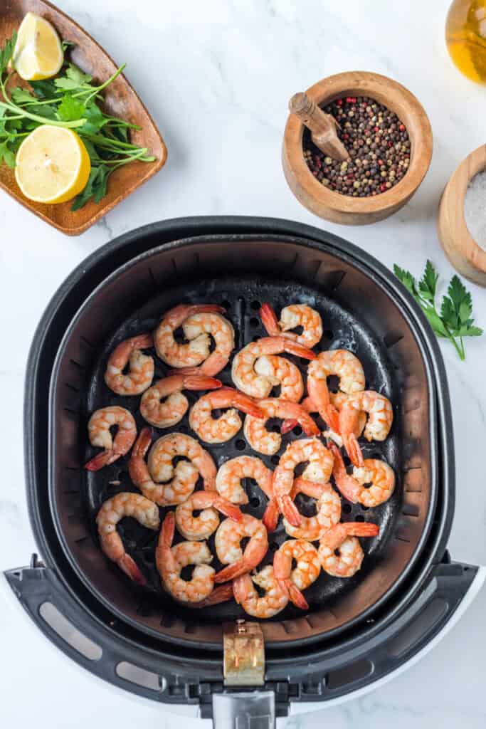 Cooked shrimp in an air fryer basket showing golden edges and pink color, surrounded by parsley, lemons, and spices.