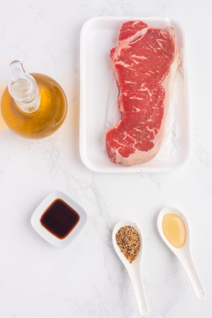 Raw strip steak with olive oil, soy sauce, honey, and Montreal steak seasoning displayed on a white surface.