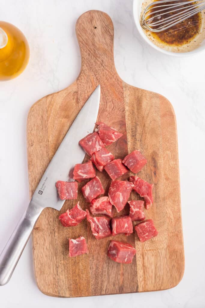 Sirloin steak cut into cubes on a wooden cutting board with a knife and marinade bowl nearby.