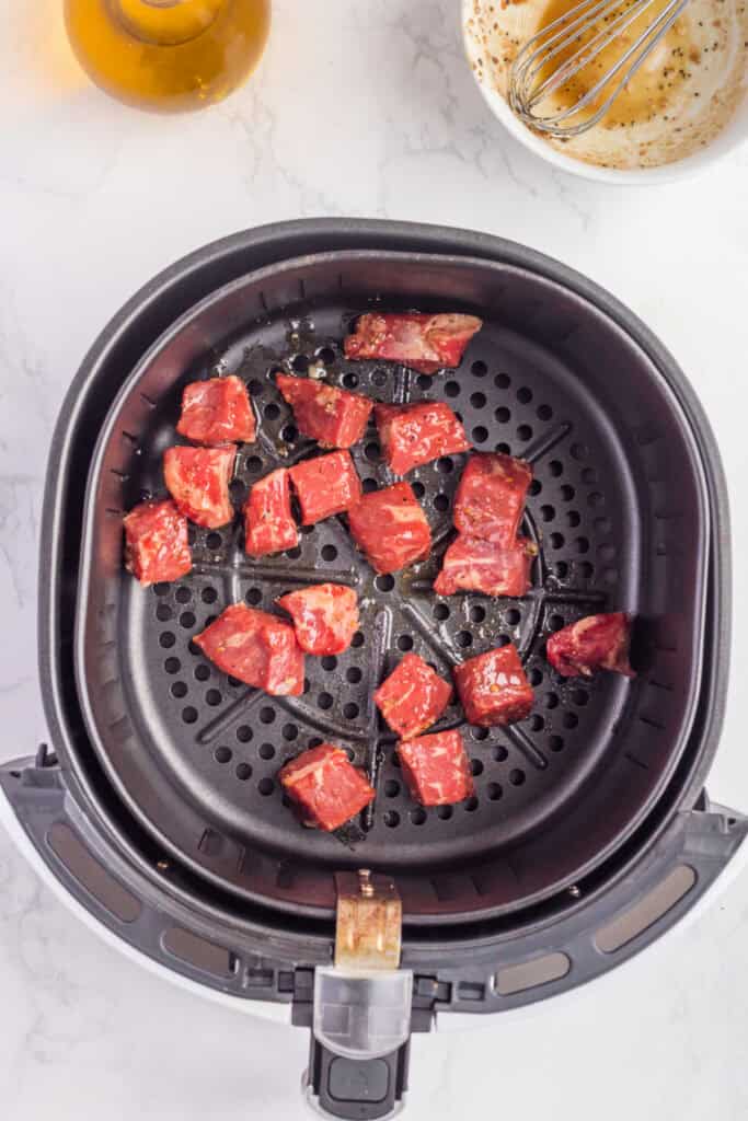 Marinated steak cubes arranged in a single layer inside an air fryer basket before cooking.
