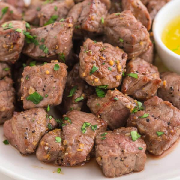 Pile of tender air fryer steak bites on a white plate with parsley garnish and garlic butter on the side.