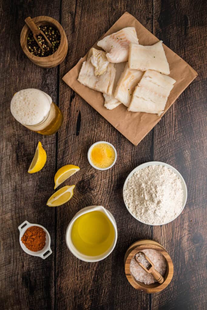 Overhead view of ingredients for beer battered fish including cod pieces, flour, beer, egg, oil, Old Bay seasoning, salt, peppercorns, and lemon wedges on a rustic wood surface.