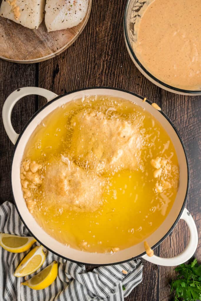 Overhead view of two battered cod pieces frying in hot oil in a white Dutch oven with bubbles forming around them.