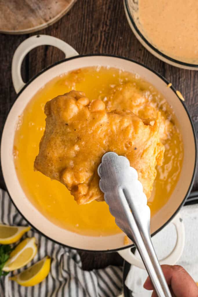 Close-up of tongs lifting a crispy golden piece of beer battered fish from hot oil.