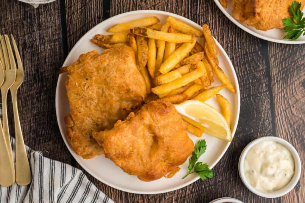 Plate of beer battered fish and fries with lemon wedge and parsley garnish, served with tartar sauce and ketchup.