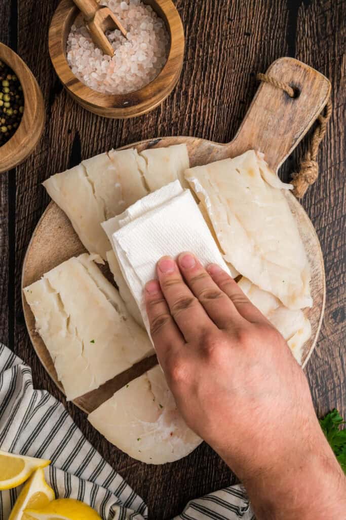 Hand patting cod filets dry with paper towels on a wooden board before seasoning for beer battered fish.