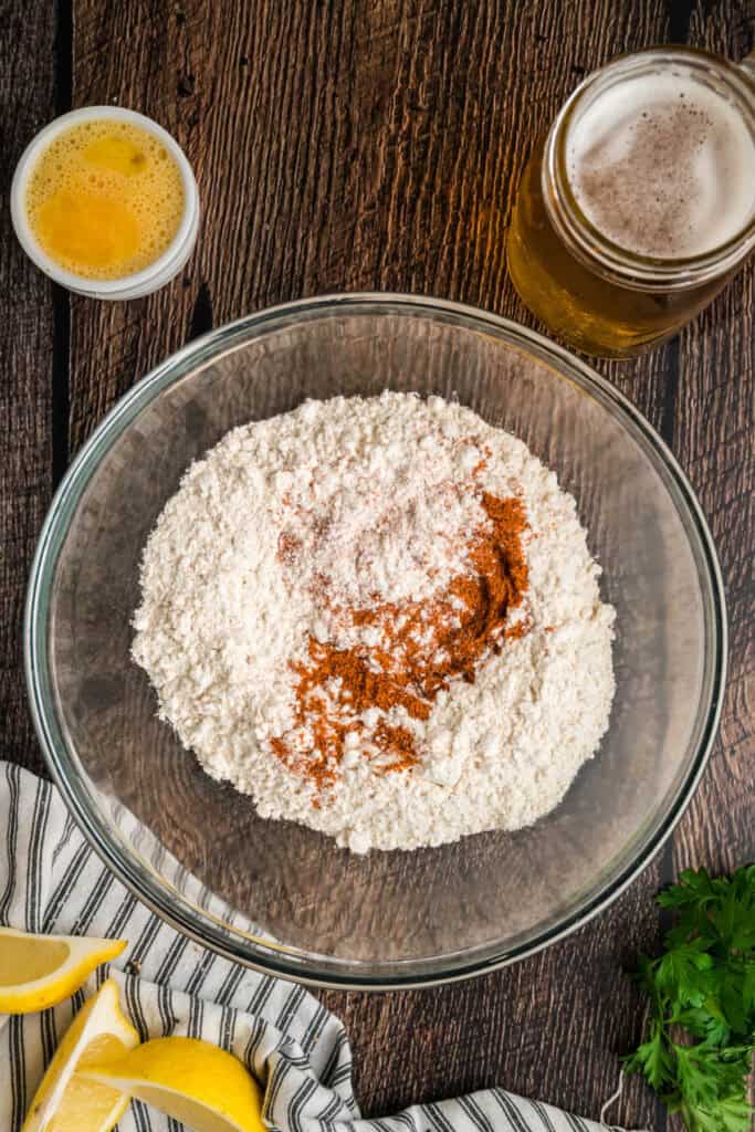 Flour and Old Bay seasoning combined in a glass mixing bowl with a jar of beer and a beaten egg nearby.