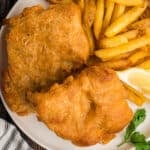 Square crop of crispy beer battered fish and fries with lemon wedge and parsley garnish on a white plate.