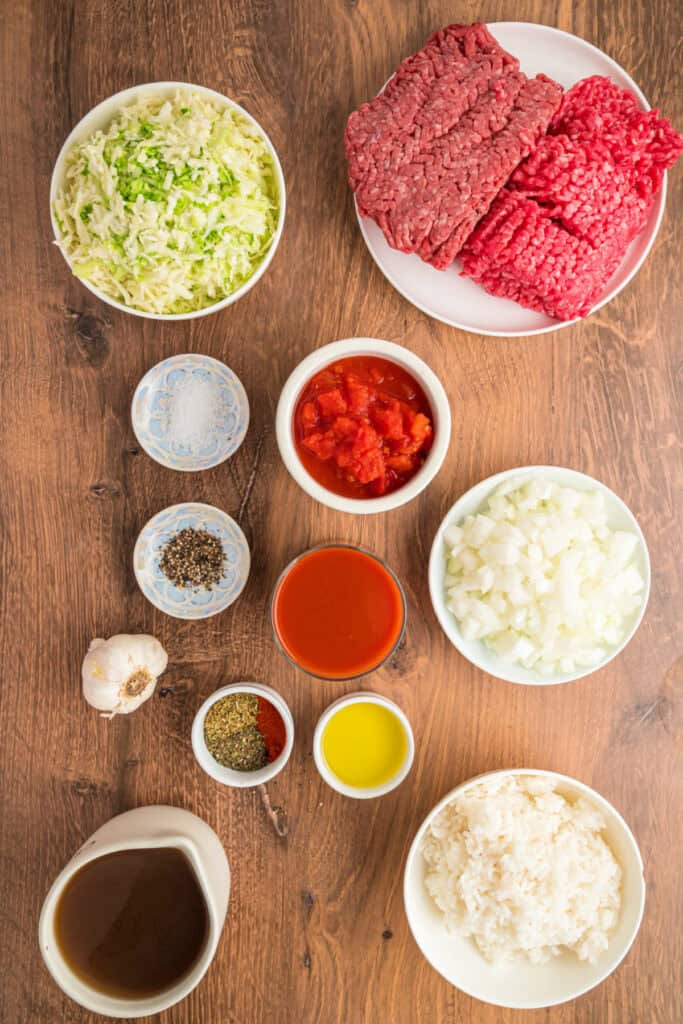 Overhead view of raw ingredients for cabbage roll soup, including ground meats, shredded cabbage, onions, tomatoes, rice, broth, and spices arranged on a wooden table.