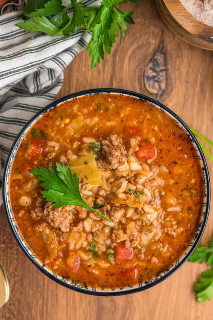 Overhead close-up of cabbage roll soup garnished with parsley in a blue-rimmed bowl on a wooden table.