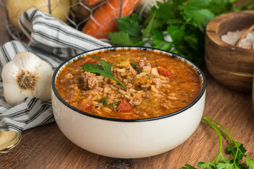 Side view of a bowl of cabbage roll soup with visible parsley garnish, garlic, and rustic kitchen props in the background.