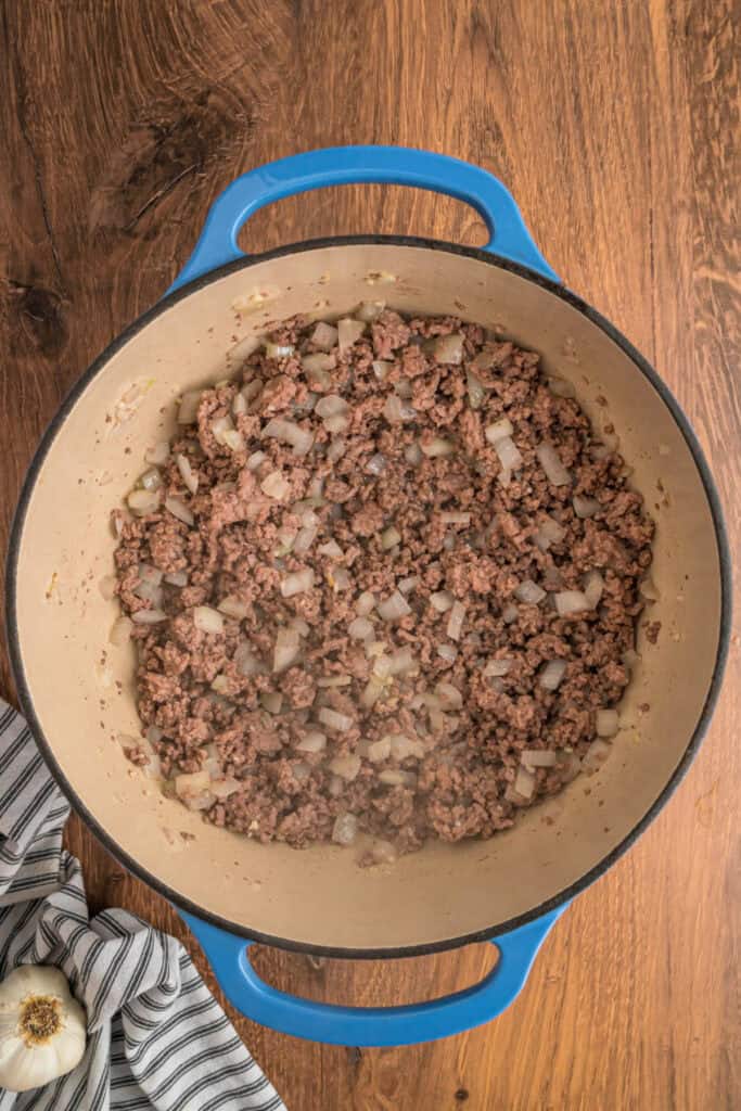 Cooked ground meat with softened onions and garlic in a blue dutch oven.