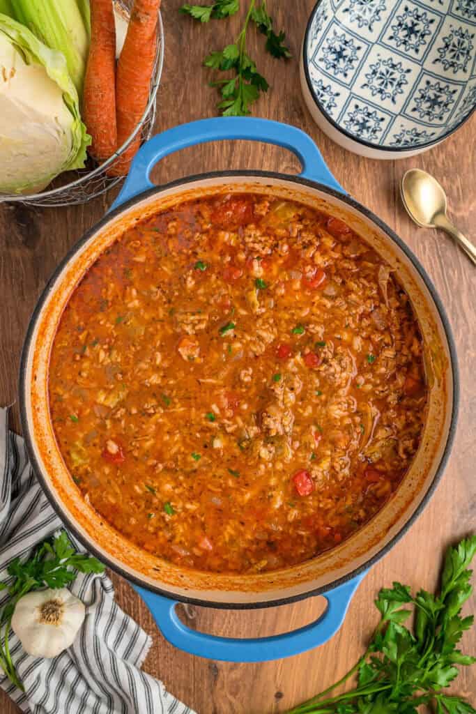 Finished cabbage roll soup simmering in a blue dutch oven surrounded by fresh parsley, cabbage, and carrots on a wooden table.