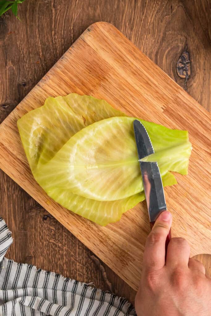 Hand using a knife to trim the thick stem from a softened cabbage leaf on a wooden cutting board.