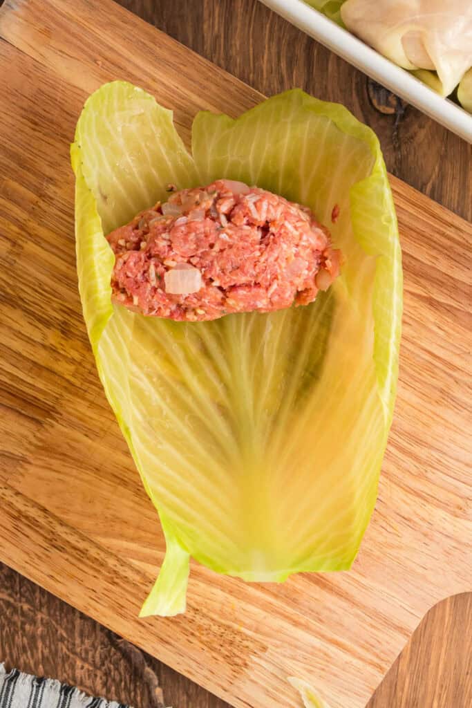 Spoonful of raw meat and rice filling placed in the center of a softened cabbage leaf on a wooden cutting board.