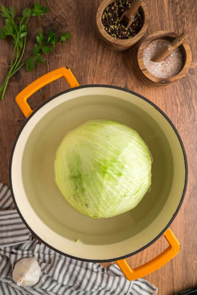 Whole green cabbage boiling in a large yellow-handled pot on a wooden surface with salt and pepper bowls nearby.