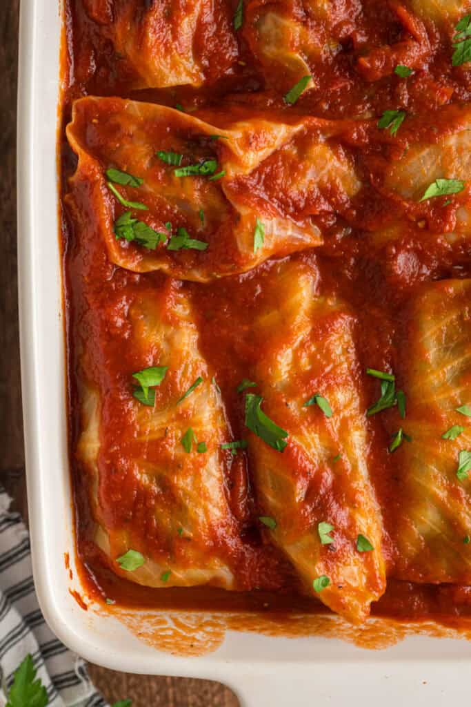 Close-up of baked cabbage rolls in a white casserole dish, covered in rich tomato sauce and sprinkled with fresh parsley.