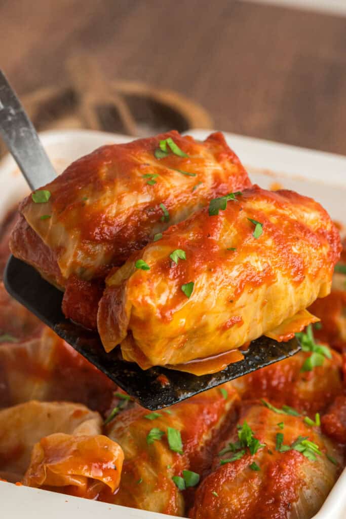 Spatula lifting two stuffed cabbage rolls from a casserole dish, coated in tomato sauce and garnished with chopped parsley.