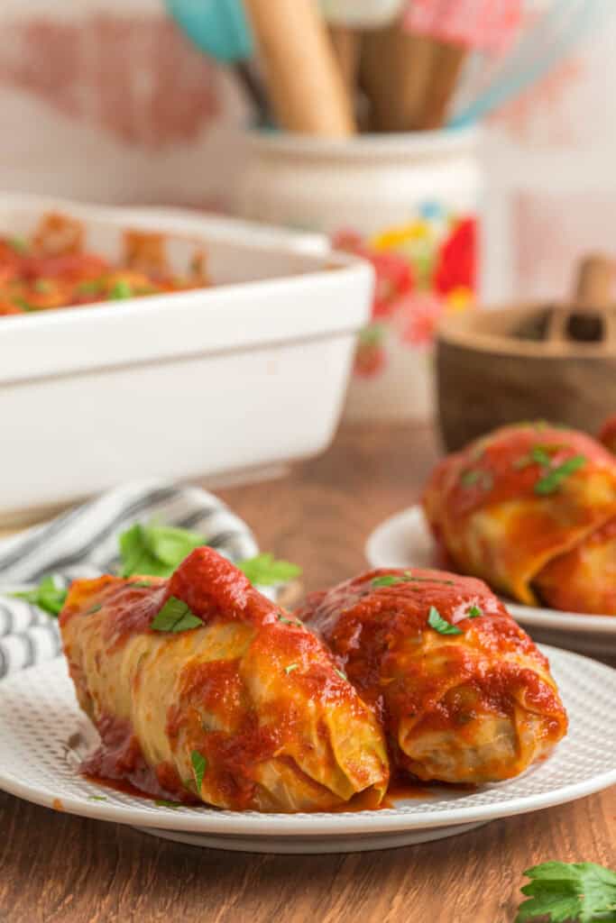 Two cabbage rolls plated with tomato sauce on a white plate, with the casserole dish blurred in the background.