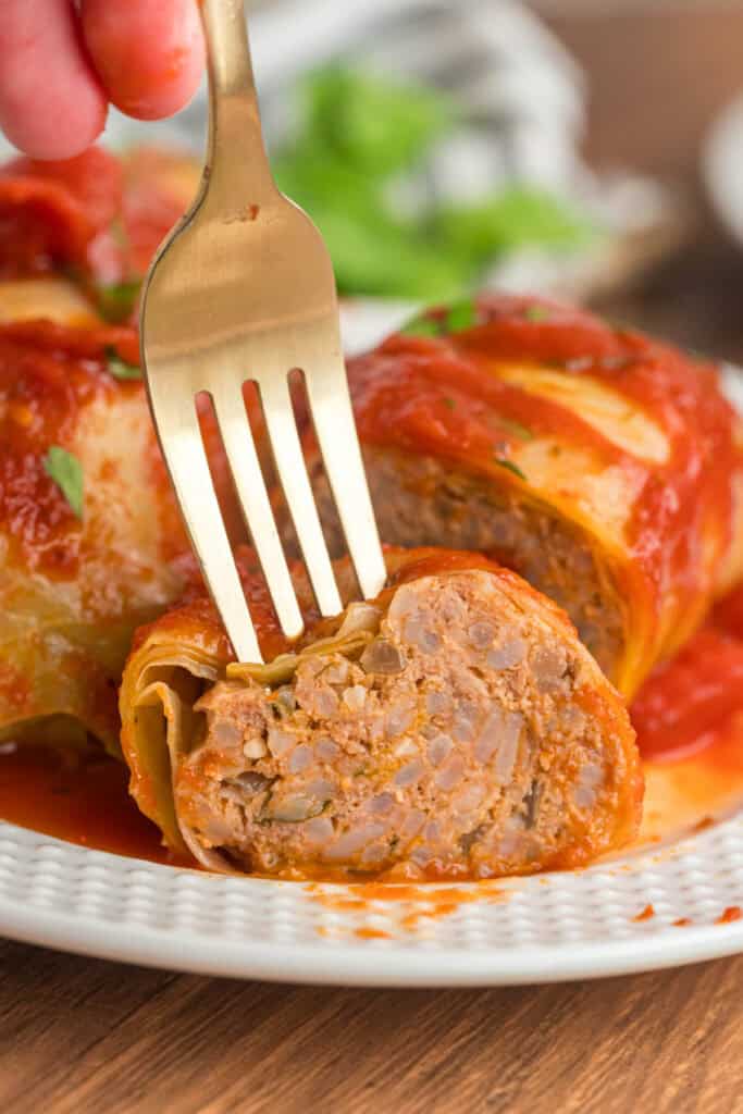 Fork cutting into a cabbage roll to reveal the beef, pork, and rice filling inside, surrounded by tomato sauce.