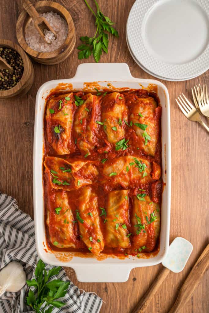 Overhead view of a white casserole dish filled with cabbage rolls in tomato sauce, on a wooden table with herbs and salt bowls.
