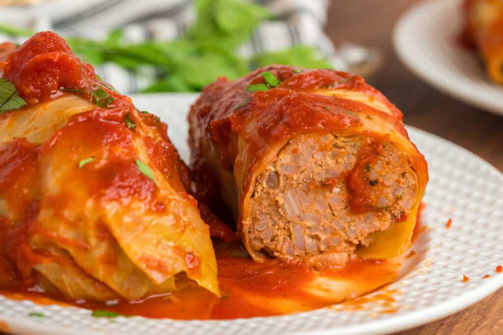 Close-up of a cabbage roll cut in half on a white plate, showing the savory beef, pork, and rice filling topped with tomato sauce.
