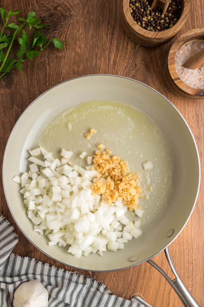 Skillet with melted butter, diced onions, and minced garlic beginning to sauté on a wooden surface.
