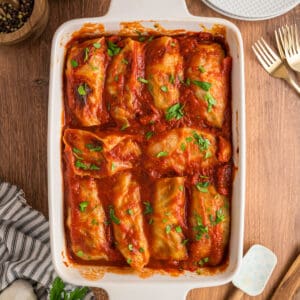 Square crop of cabbage rolls baked in tomato sauce, topped with parsley in a white baking dish on a rustic wooden table.