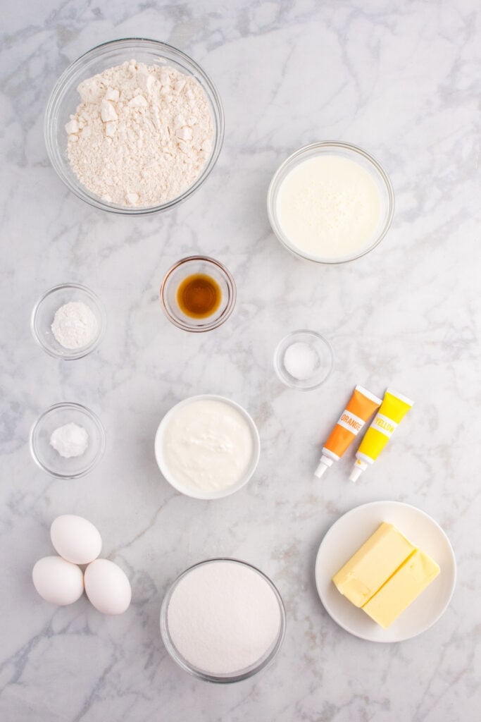 Overhead view of measured cupcake ingredients including flour, sugar, butter, eggs, yogurt, buttermilk, and food coloring on a marble surface.