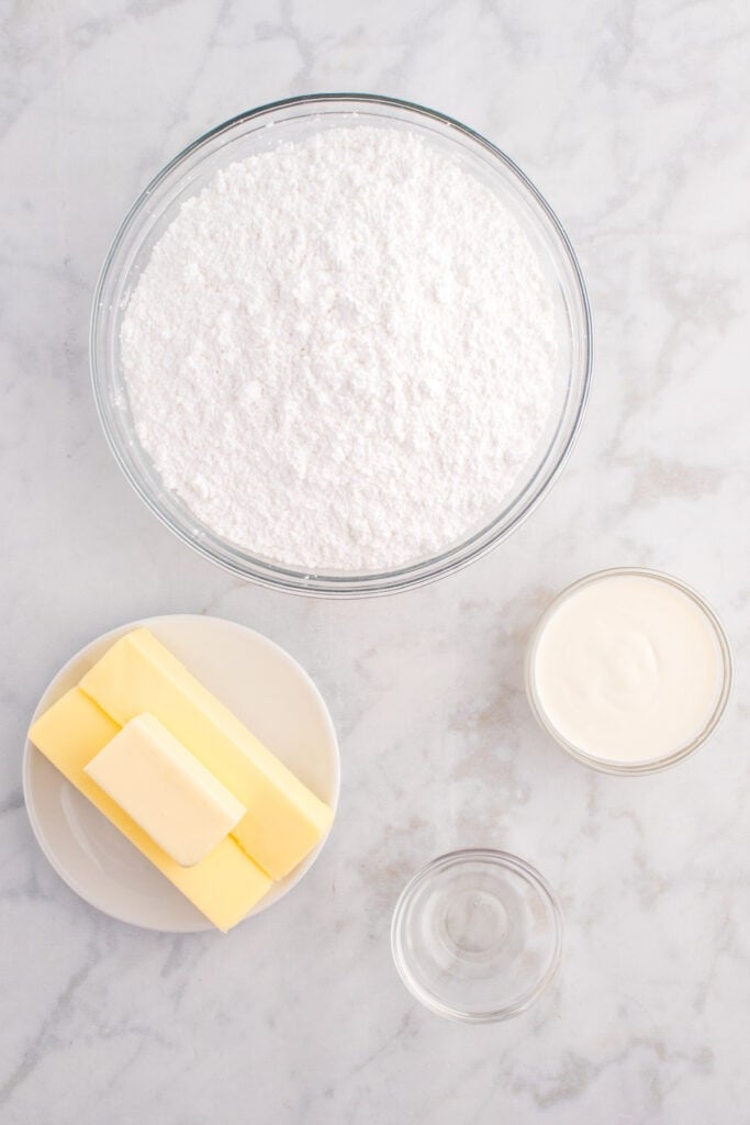 Overhead view of frosting ingredients including powdered sugar, butter, cream, and clear vanilla extract in glass bowls.