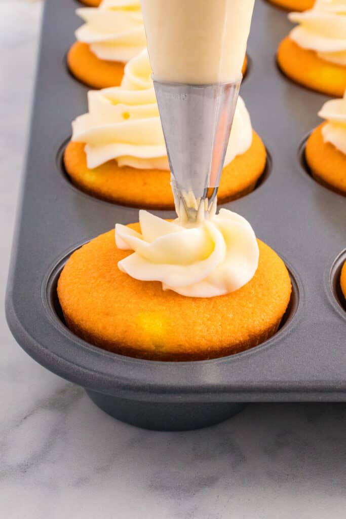 Piping bag swirling white frosting onto cooled orange cupcakes in a muffin pan.
