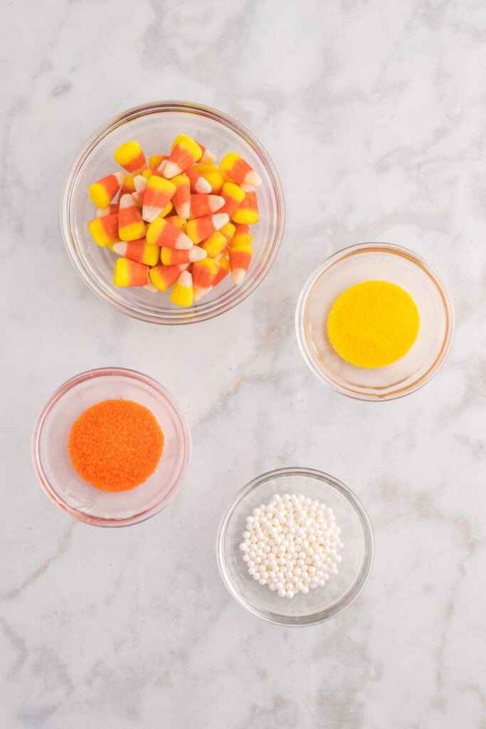 Small glass bowls filled with candy corn, orange sanding sugar, yellow sanding sugar, and white sugar pearls for decorating.