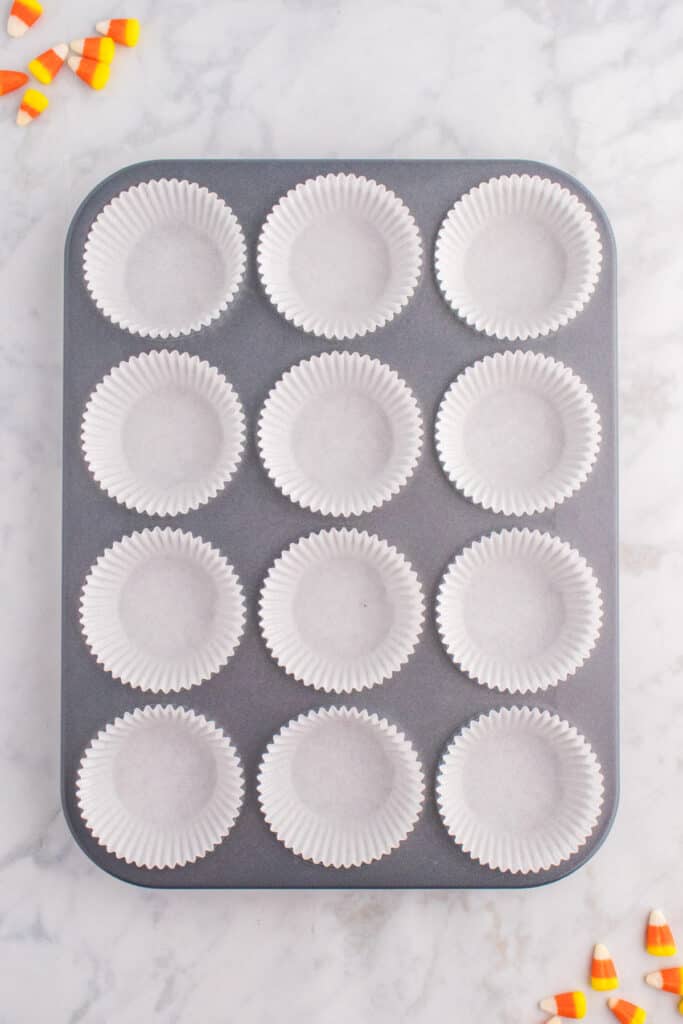 Muffin pan lined with white cupcake liners and candy corn scattered around on marble surface.