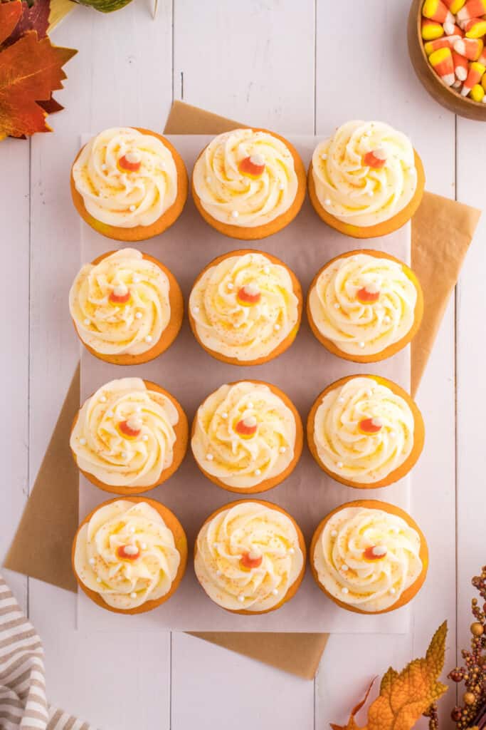 Overhead view of frosted Candy Corn Cupcakes arranged neatly on parchment with fall leaves and candy corn accents.