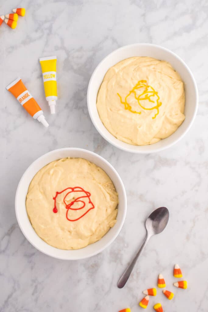 Two bowls of cupcake batter with yellow and orange gel food coloring drizzled on top before stirring.